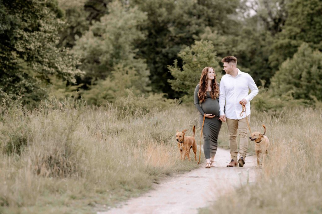 Paar geht über einen Weg in der Natur. Die Frau ist schwanger und schaut ihren Mann verliebt an. Links und rechts laufen beide Hunde des Paares.