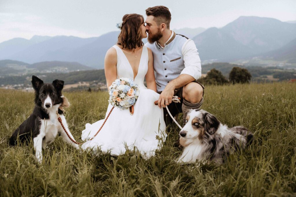 Hochzeitspaar kniet vor Bergkulisse und küsst sich. Ihre beiden Hunde - ein Border Collie und ein Australian Shepherd liegen links und rechts von ihnen im Gras