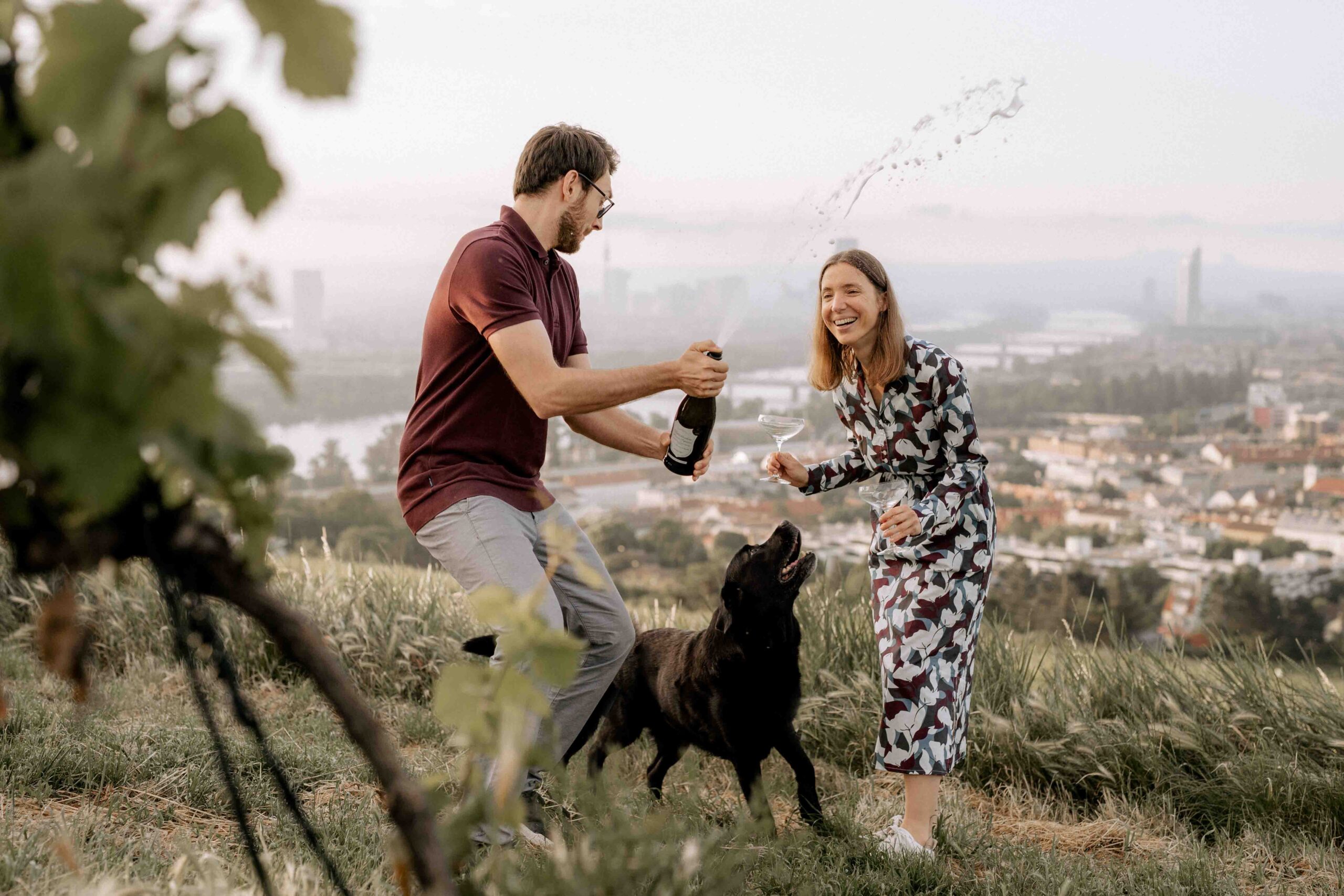 Hundefotoshooting Nussberg Wien. Mensch und Hund stehen auf Berg mit der Stadt im Hintergrund und lassen die Korken einer Sektflasche knallen.