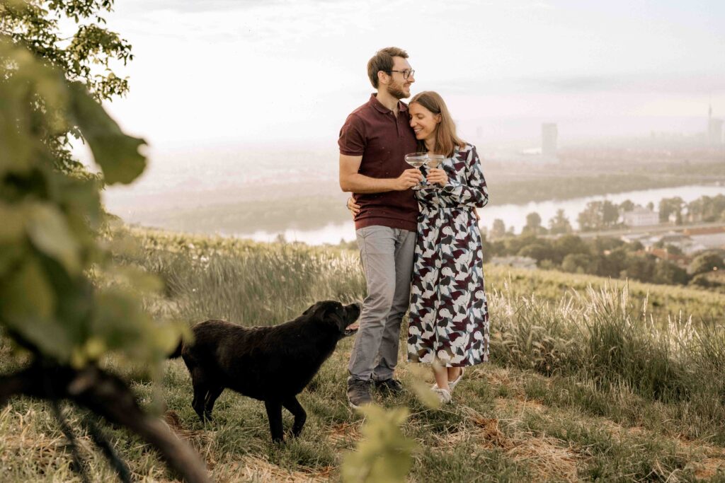 Paar kuschelt sich im stehen aneinander und hält dabei zwei Weingläser in der Hand. Im Hintergrund sieht man die Stadt Wien, im Vordergrund erkennt man Weinreben. Vor dem Paar steht noch ihr schwarzer Labrador, der sie liebevoll ansieht.